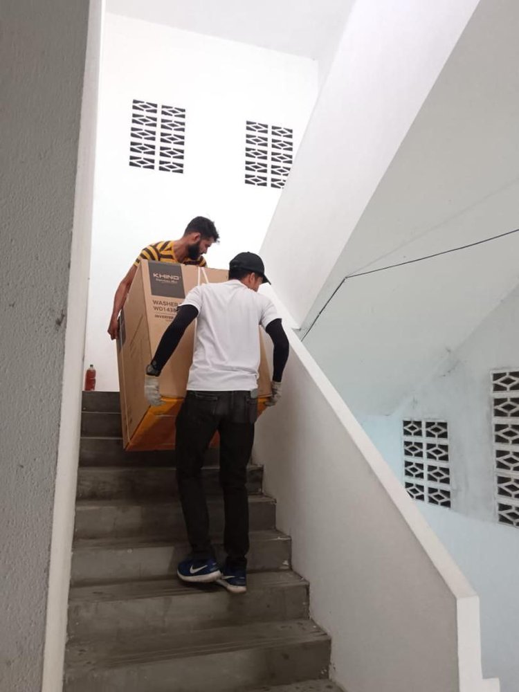 Dua petugas mengangkat kotak washer dryer menaiki tangga